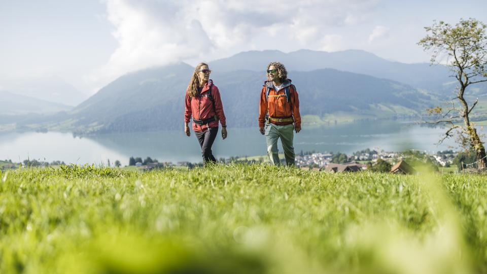 Eine Frau und ein Mann wandern auf einer Wiese hoch über einem Dorf. Im Hintergrund sind ein See und bewaldete Hügel zu sehen.