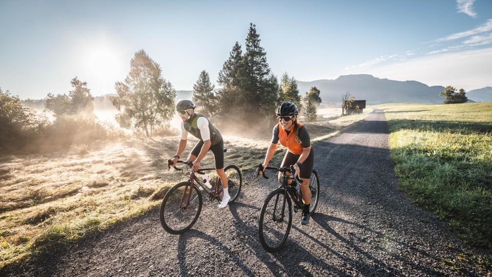 Ein paar fährt auf dem Gravelbike auf einer Kiesstrasse in der Morgensonne.