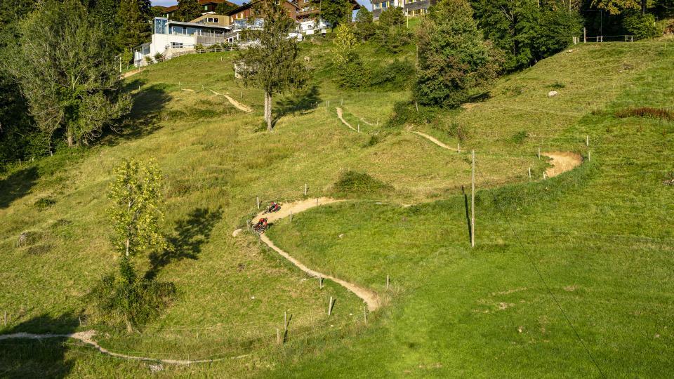 Zwei Mountainbikende fahren auf der gebauten Strecke in eine Anlegerkurve. Der Trail schlängelt sich durch grüne Wiesen.