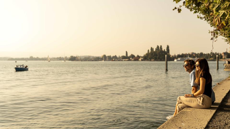 Zwei Menschen sitzen am Ufer des Zugersee und blicken in den goldgelben Sonnenuntergang.