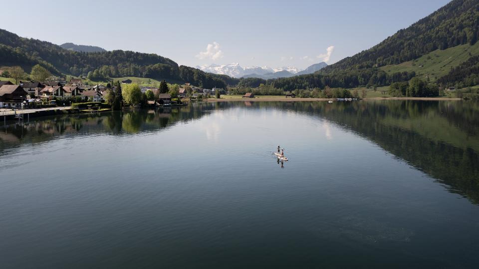 Zwei Stand-up-Paddelnde sind auf dem Ägerisee. Im Hintergrund sind die schneedeckten Gipfel der Alpen zu sehen.