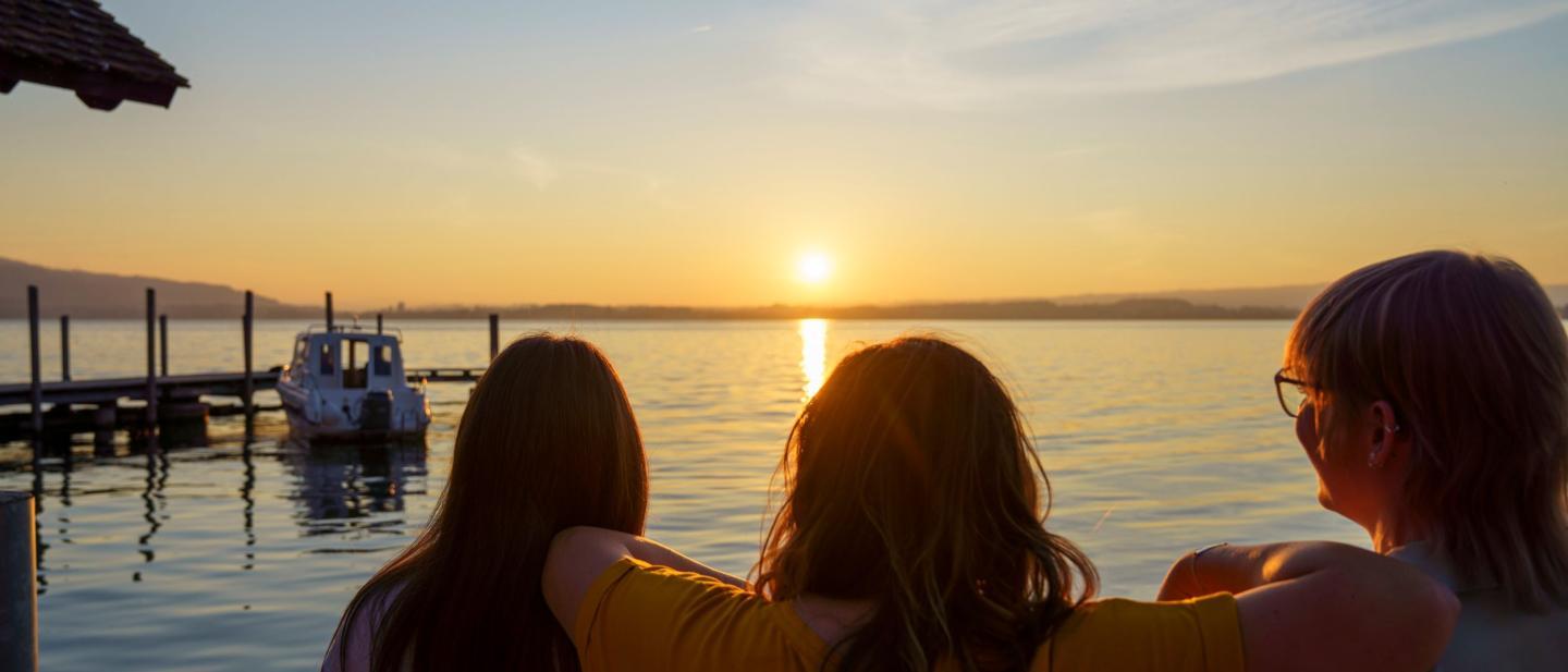 Drei Frauen sitzen am Ufer des Zugersee und schauen in den Sonnenuntergang