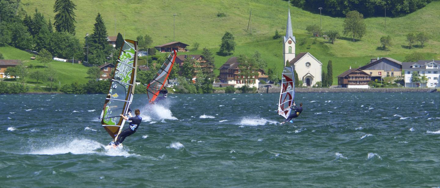 Drei Windsurfer surfen auf dem unruhigen Ägerisee. Im Hintergrund sind die Kirche und Holzhäuser zu sehen.