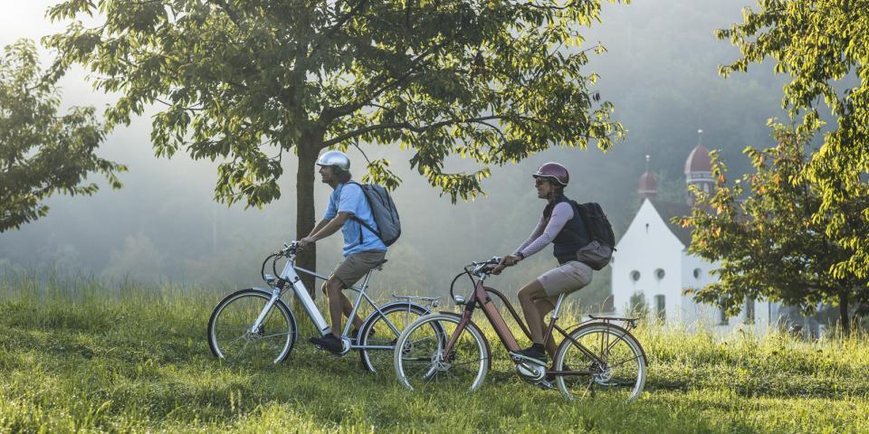 Eine Frau und eine Mann fahren auf dem Velo vor einer Kapelle durch. Die Kapelle steht auf einer Wiese und ist mit Kirschbäumen umgeben.